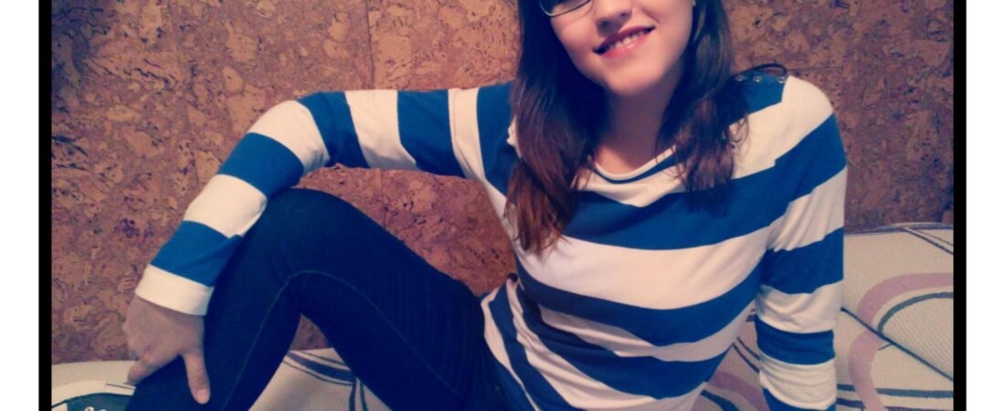 Joven con gafas y camiseta de rayas anchas azules y blancas sentada ante una pared de corcho