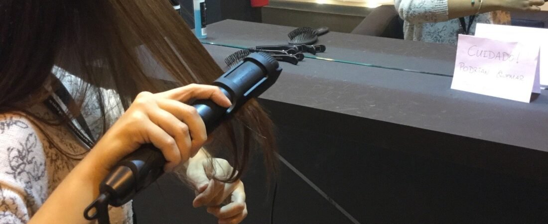 Mujer con gafas rizándose el pelo con unas tenacillas ante el espejo de un estand de peluquería