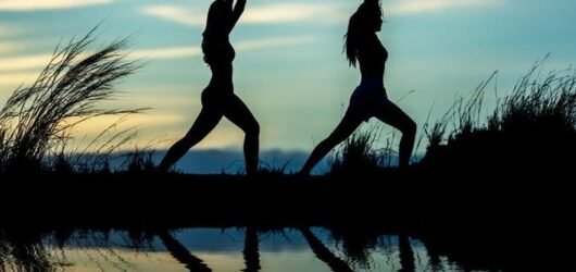 Dos siluetas practicando yoga a la orilla de un lago al atardecer, con su reflejo en el agua