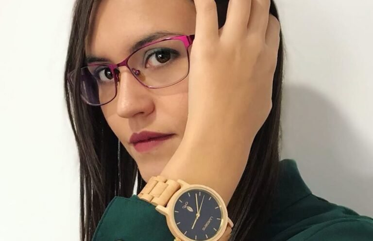 Mujer con gafas fucsia y camisa verde mostrando un reloj de madera de esfera azul