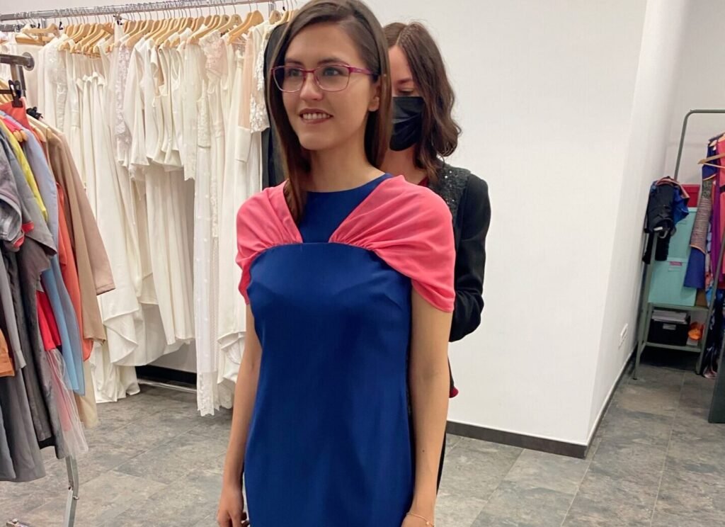 Mujer con gafas probándose un vestido azul con pañuelo rosa mientras una dependienta se lo ajusta